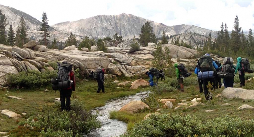 A group of backpackers hike through a grassy area with large rock formations and a creek. 